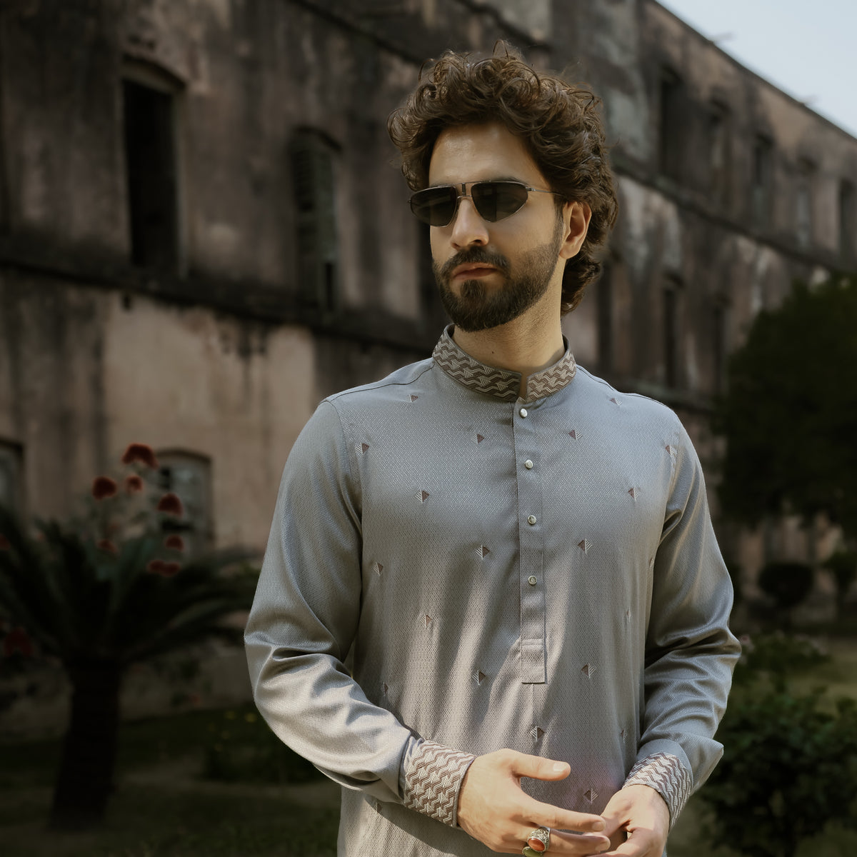A man wearing a light gray embroidered kurta and sunglasses, standing outdoors in front of a weathered building.