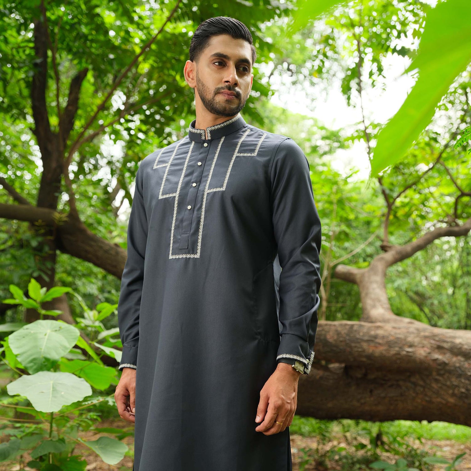 Man wearing a dark traditional outfit standing in a lush green forest.
