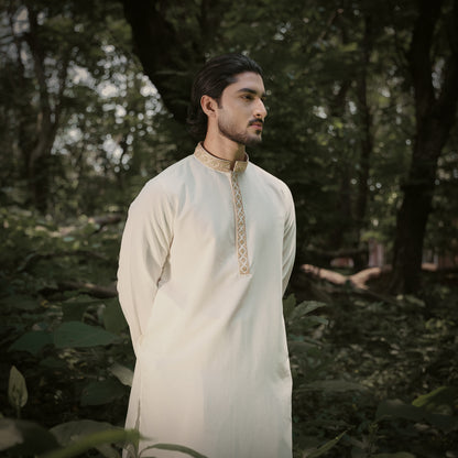 A man wearing a cream-colored traditional YAMAAN panjabi with gold embroidery on the collar and front placket, standing in a lush, green forest.