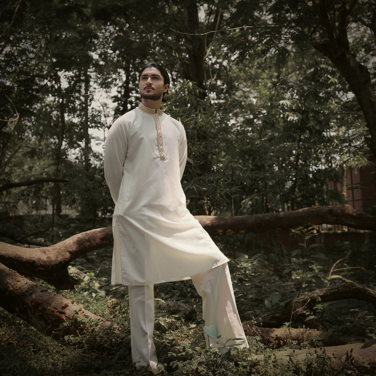 A man wearing a white YAMAAN traditional panjabi and trousers stands outdoors in a wooded area with fallen logs and trees in the background.
