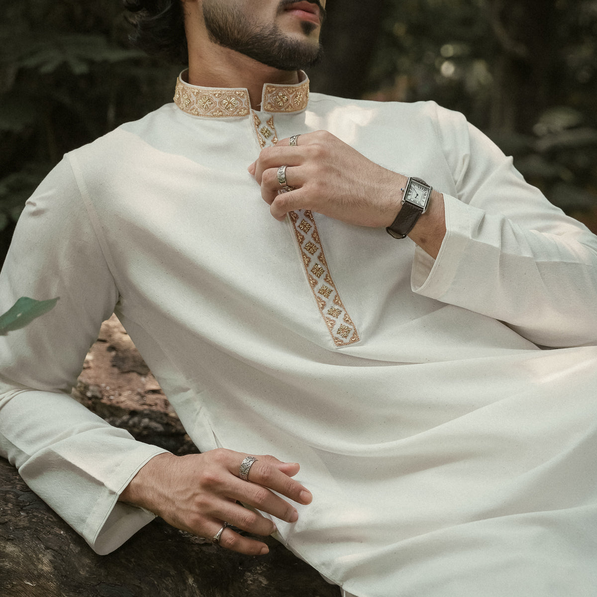 A man wearing a white long-sleeve YAMAAN panjabi with gold embroidered detailing on the collar and chest, posed outdoors against a blurred green background.