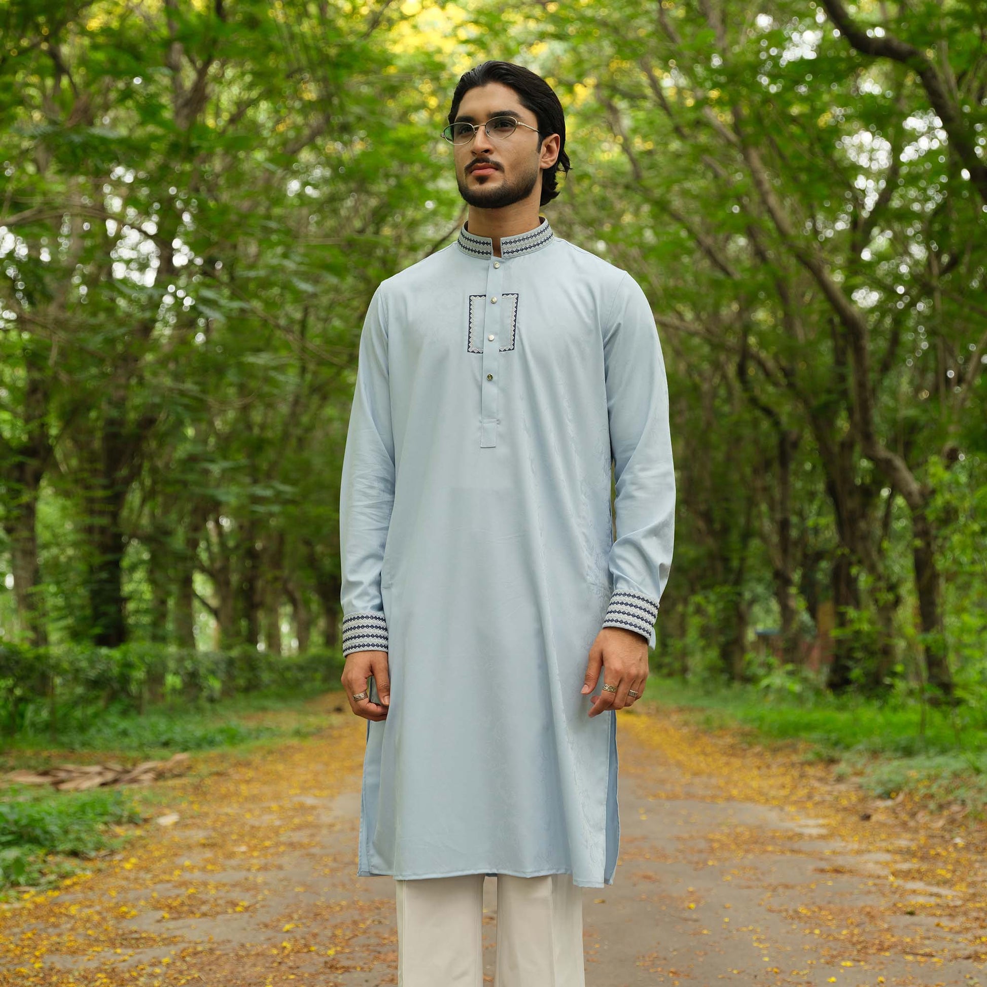 Man wearing a light gray kurta with white pants standing in a forest.
