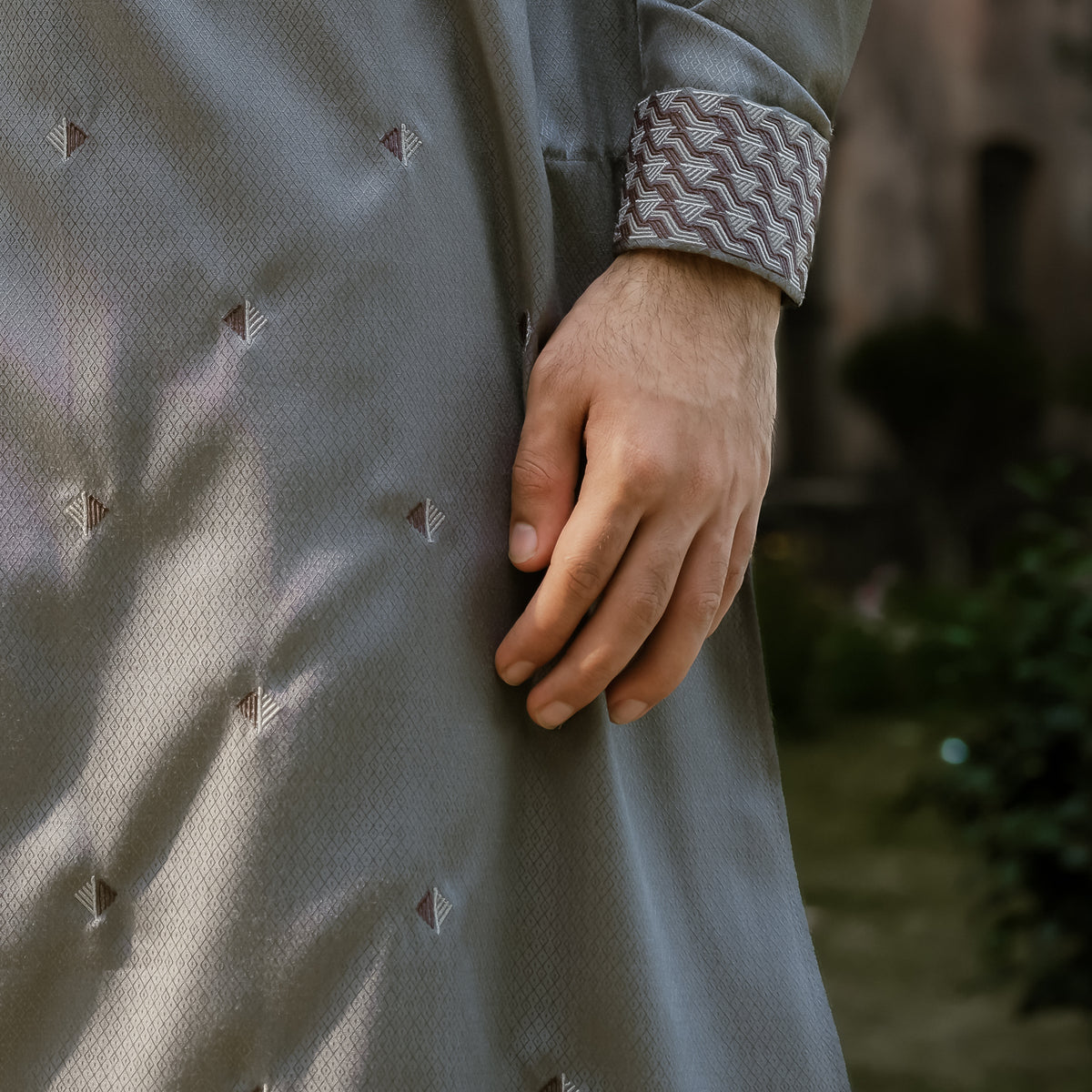 Close-up of a person wearing a light gray textured YAMAAN PANJABI or tunic with small geometric embroidered details and a patterned cuff, photographed outdoors with a blurred background.