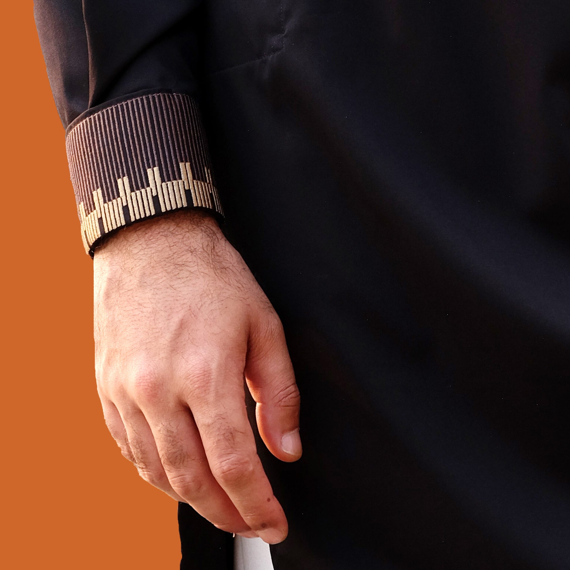 YAMAAAN Close-up of a person's hand wearing a dark coat with a patterned cuff against an orange background
