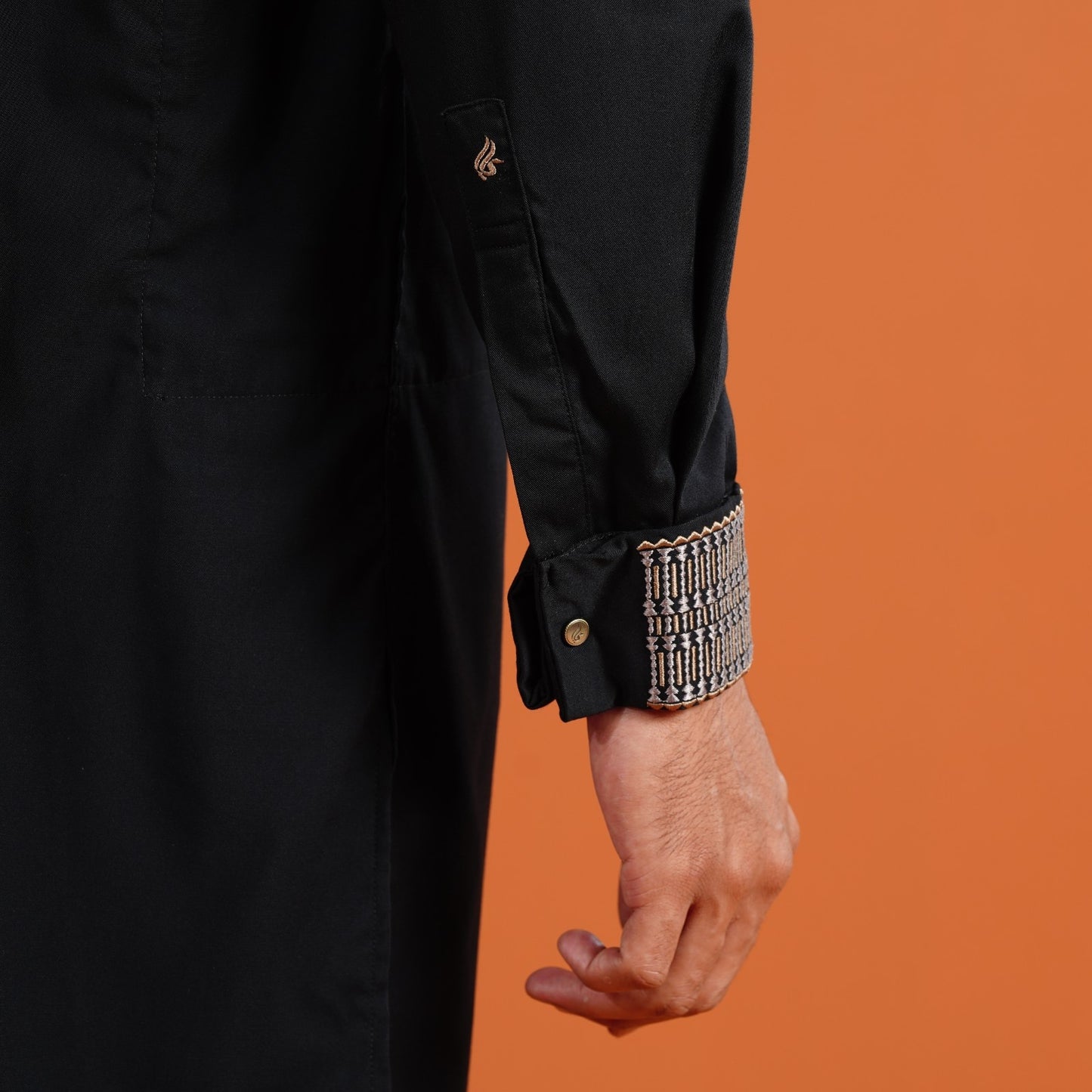 YAMAAN 
Close-up of a person wearing a black coat with an ornate cuff against an orange background