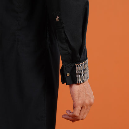 YAMAAN 
Close-up of a person wearing a black coat with an ornate cuff against an orange background