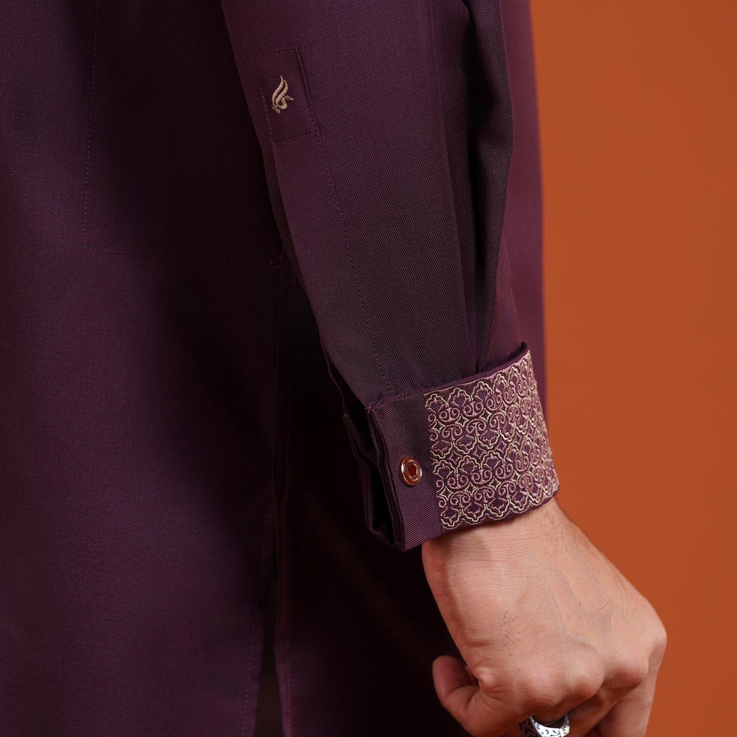 YAMAAN Close-up of a person wearing a dark maroon garment with intricate cuff design on an orange background