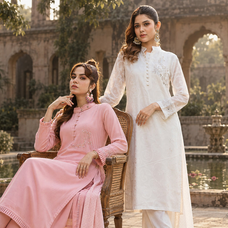 Two women modeling traditional YAMAAN women's wear in an outdoor courtyard setting. One is wearing a light pink long-sleeve kameez with silver embroidery, seated on a wicker chair, and the other is wearing a white long-sleeve kameez with floral embroidery, standing beside the chair.