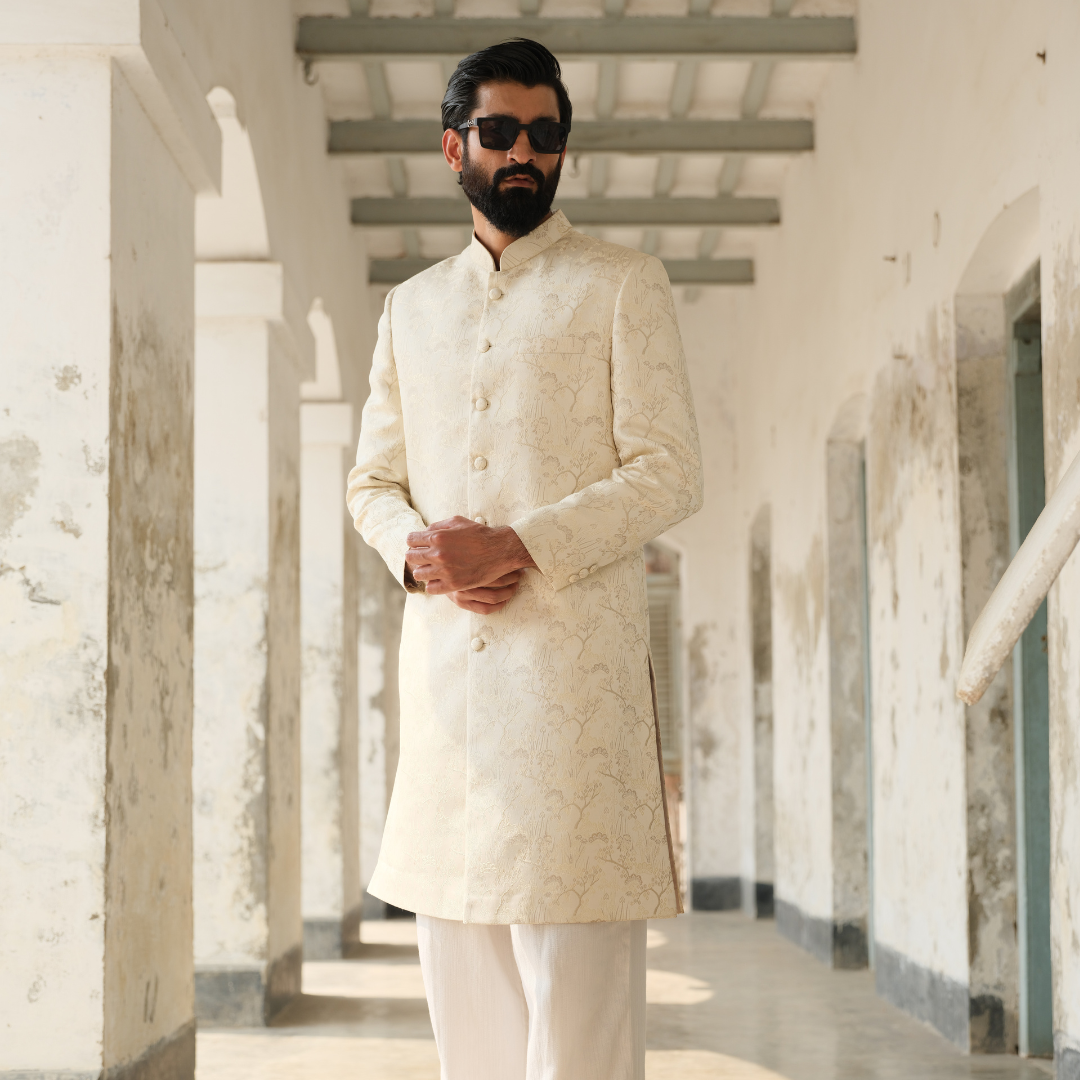 Man wearing a cream sherwani in a hallway with white walls and columns.
