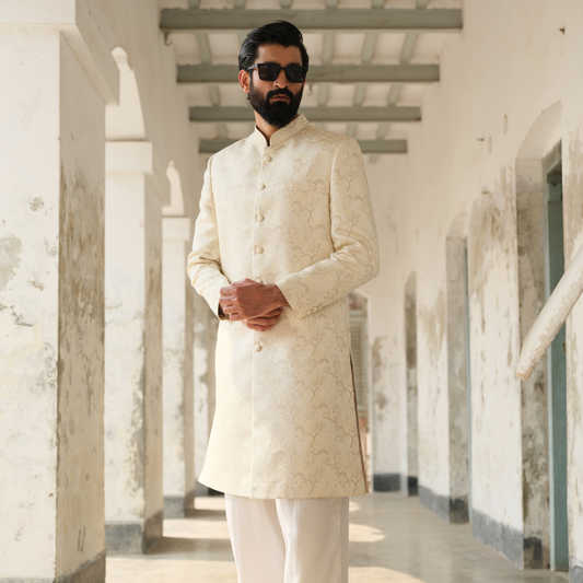 Man wearing a cream sherwani in a hallway with white walls and columns.