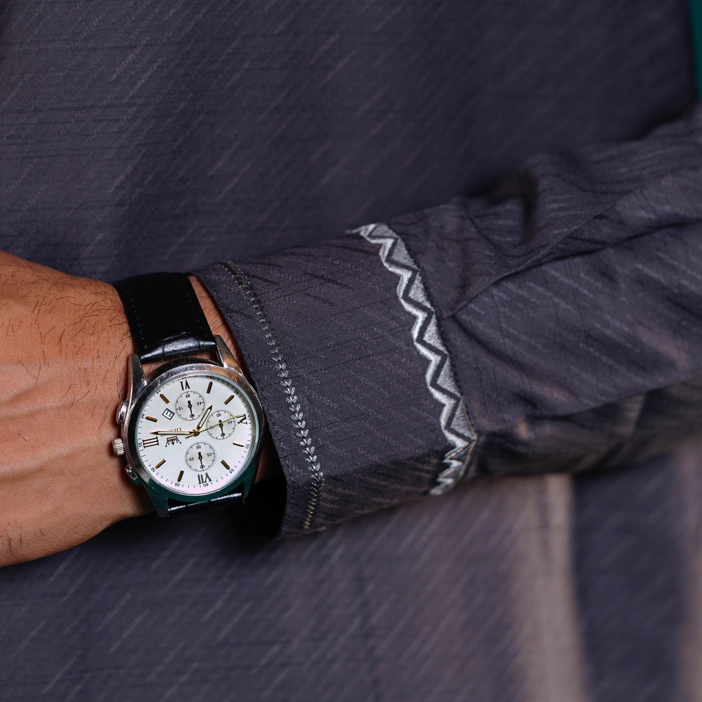Yamaan grey panjabi close-up of a wrist wearing a watch with a dark shirt sleeve.